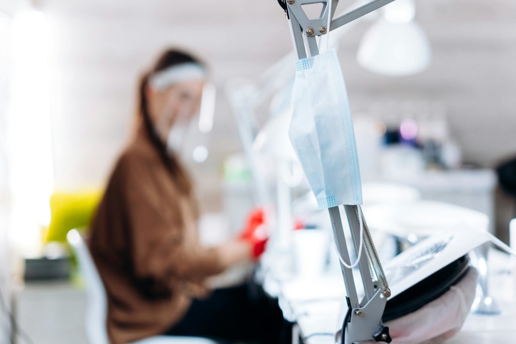Focus on a face mask hanging in a blurred office with a person working, symbolizing the new normal.